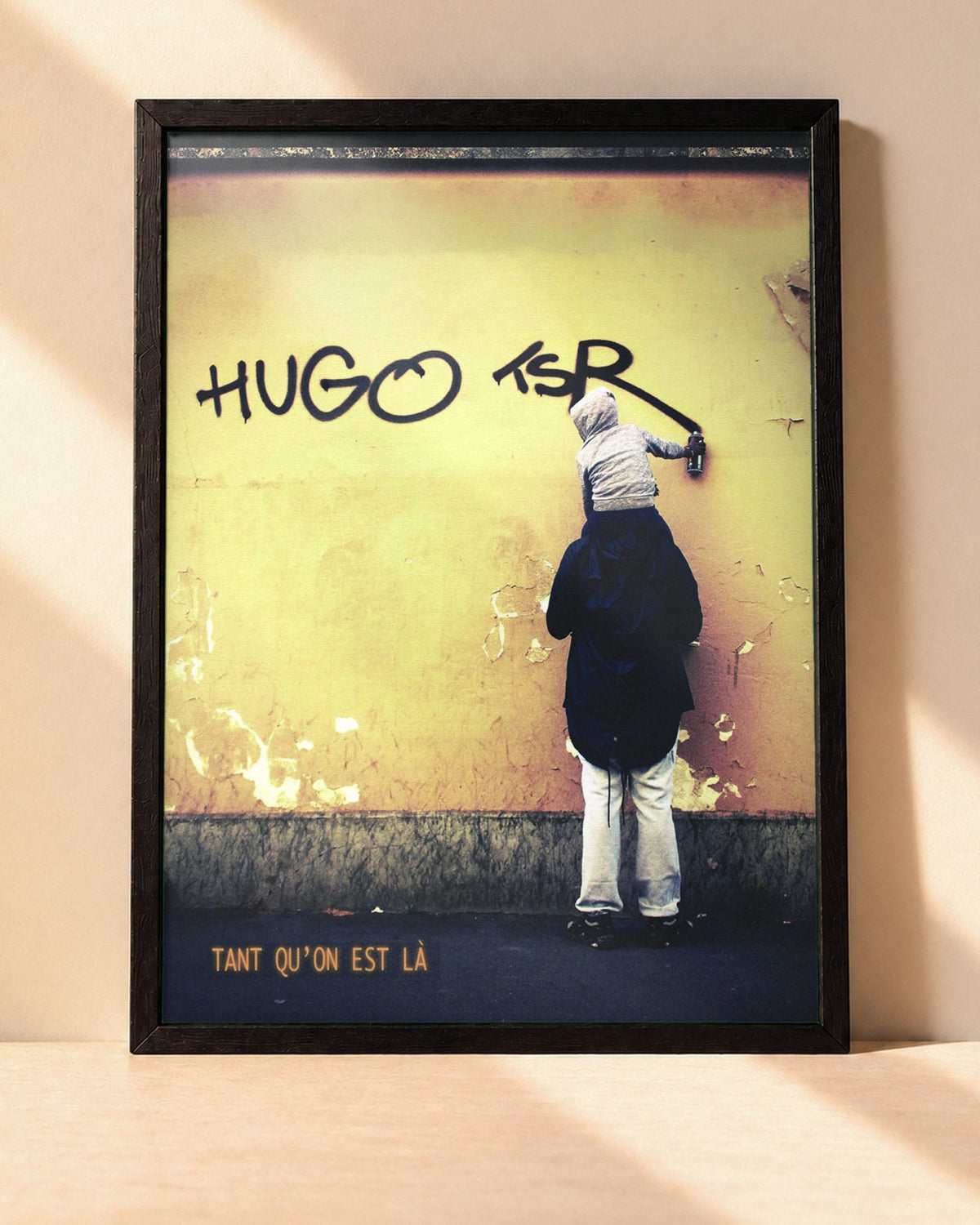 Poster Hugo TSR – Tant Qu'on Est Là encadré dans un cadre noir, posé contre un mur clair