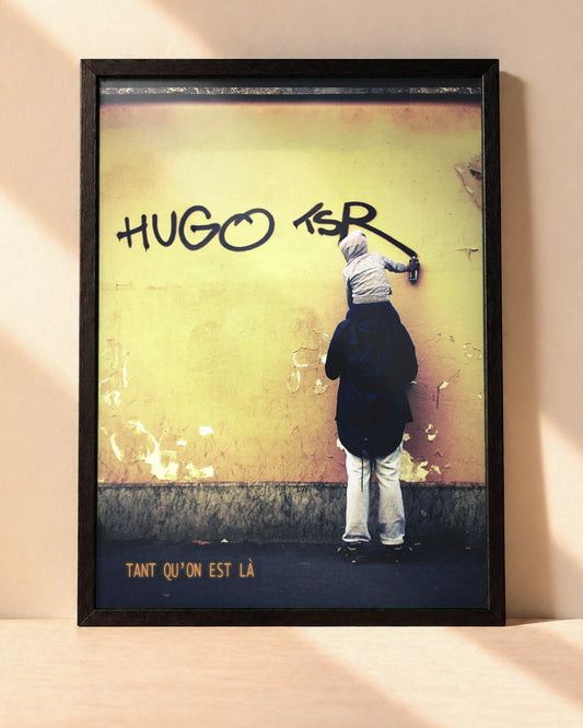 Poster Hugo TSR – Tant Qu'on Est Là encadré dans un cadre noir, posé contre un mur clair